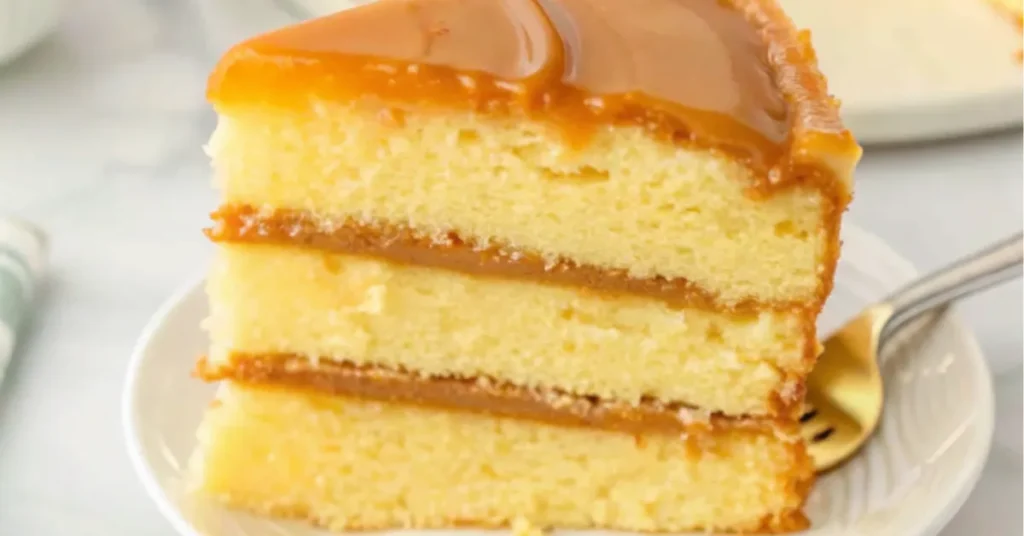 A slice of classic Southern caramel cake with rich caramel frosting between three moist vanilla cake layers on a white plate.